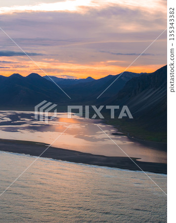 Aerial view of Iceland's Stokknes Peninsula showing calm ocean waters, a black sand beach, rugged mountains, and a vibrant orange and purple sunset. 135343382