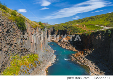 Studlagil Canyon in Iceland features basalt columns, a turquoise river, green hills, and a viewing platform under a clear blue sky with scattered clouds. Studlagil Canyon in Iceland features basalt columns, a turquoise river, green hills, and a viewing platform under a clear blue sky with scattered clouds. 135343387