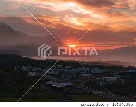 A vibrant sunset illuminates an Icelandic village surrounded by rugged terrain. Misty mountains rise in the background, adding depth to the scene. A vibrant sunset illuminates an Icelandic village surrounded by rugged terrain. Misty mountains rise in the background, adding depth to the scene. 135343390