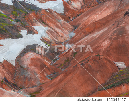 Vibrant red, orange, and brown rhyolite mountains with white snow patches and rising steam from geothermal vents in Iceland's highlands. 135343427