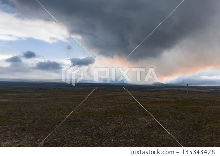 Expansive barren terrain with sparse vegetation and rocky ground under dark clouds. Orange and pink hues suggest sunset or volcanic activity. Steam rises in the distance. 135343428