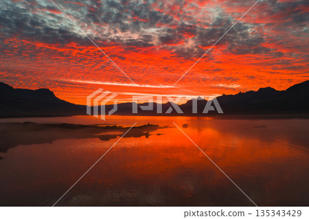 A vivid sunset in Iceland reflects red and orange hues on calm water, with distant mountain silhouettes and unique cloud formations in the sky. A vivid sunset in Iceland reflects red and orange hues on calm water, with distant mountain silhouettes and unique cloud formations in the sky. 135343429