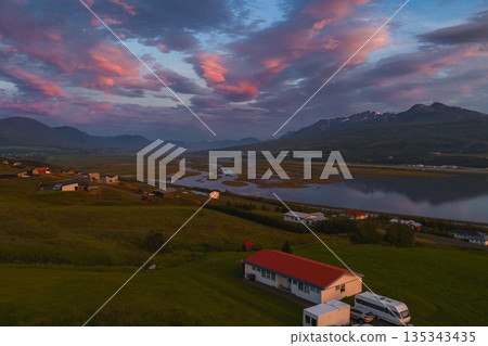 Serene aerial view of a rural Icelandic landscape at sunset. A calm river reflects pink and orange hues, with houses and green fields in the foreground. Serene aerial view of a rural Icelandic landscape at sunset. A calm river reflects pink and orange hues, with houses and green fields in the foreground. 135343435