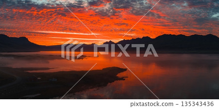 Vivid red and orange sunset reflects on still waters, with silhouetted peaks and mist. A cluster of buildings sits near the shoreline in the foreground. Vivid red and orange sunset reflects on still waters, with silhouetted peaks and mist. A cluster of buildings sits near the shoreline in the foreground. 135343436