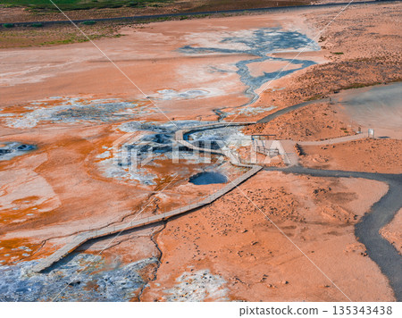 Geothermal area in Iceland with orange and white mineral deposits, steaming vents, bubbling mud pots, and a wooden boardwalk winding through the scene. 135343438