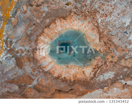 Aerial view of a geothermal Strokkur Geyser in Iceland featuring a vivid blue pool surrounded by orange, brown, and white mineral deposits and textured terrain. Aerial view of a geothermal Strokkur Geyser in Iceland featuring a vivid blue pool surrounded by orange, brown, and white mineral deposits and textured terrain. 135343452