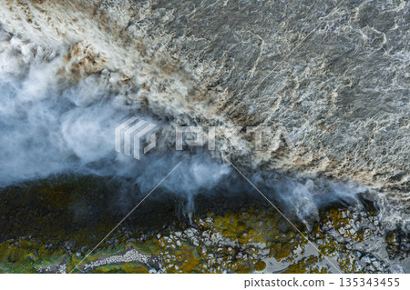 Aerial perspective of Dettifoss waterfall in Iceland, showcasing cascading water, mist plumes, moss covered rocks, and rugged earthy terrain. 135343455