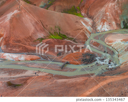Aerial view of Iceland's Rainbow Mountains with vibrant red, orange, and green hues. A river with a wooden footbridge and small figures are visible. Aerial view of Iceland's Rainbow Mountains with vibrant red, orange, and green hues. A river with a wooden footbridge and small figures are visible. 135343467