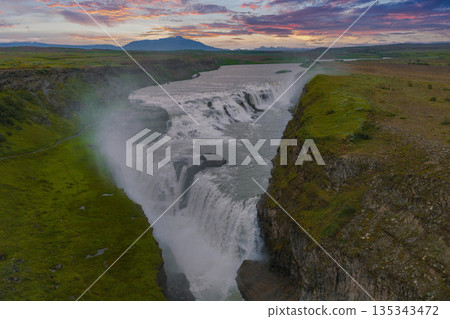 Gullfoss waterfall in Iceland cascades in two stages into a canyon, surrounded by green terrain, distant mountains, and a colorful sunset sky. 135343472