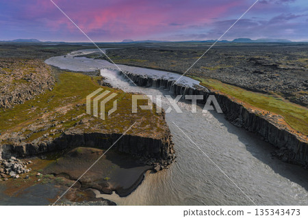 Studlagil Canyon in Iceland features basalt columns, a winding river, and vibrant moss under a pink and purple sky during sunset or sunrise. 135343473