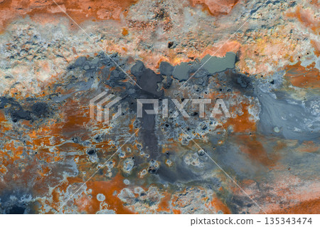 Aerial view of a geothermal area in Iceland featuring vibrant orange, red, and gray hues, bubbling mud pots, steaming vents, and mineral deposits. 135343474