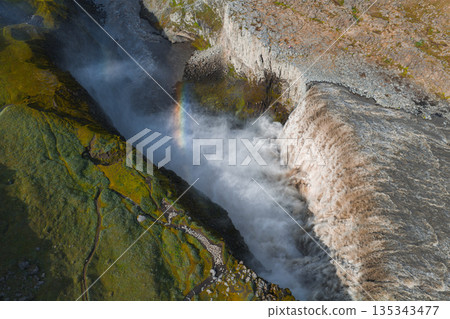 Aerial view of Dettifoss waterfall in Iceland, showcasing cascading water, mist, a vivid rainbow, rugged cliffs, and lush green moss. 135343477