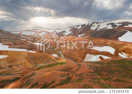Vibrant rhyolite hills of Iceland's Rainbow Mountains with snow patches, a winding stream, and sun rays breaking through a dramatic cloudy sky. 135343478