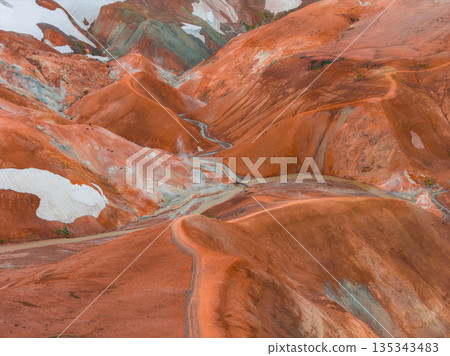 Vibrant multicolored rhyolite hills of Iceland's Rainbow Mountains, featuring red, orange, and green hues with snow patches and a winding trail. 135343483