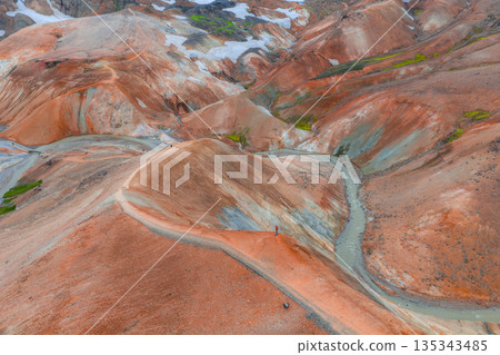 Vibrant rhyolite hills in Landmannalaugar, Iceland, with orange, red, and brown hues, green moss, snow patches, a winding trail, and a small stream. 135343485