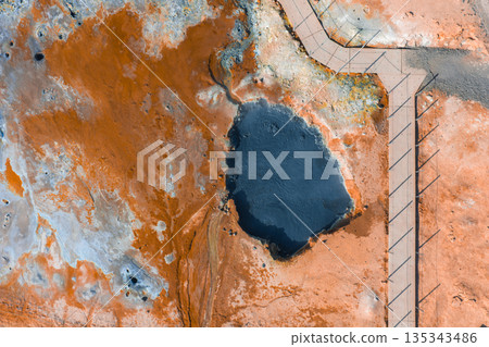 An aerial view of a geothermal area in Iceland featuring orange and rust colored terrain, a steaming hot spring, and a wooden boardwalk with railings. An aerial view of a geothermal area in Iceland featuring orange and rust colored terrain, a steaming hot spring, and a wooden boardwalk with railings. 135343486