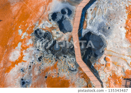 Aerial image of a geothermal area in Iceland featuring orange and white mineral deposits, steaming fumaroles, bubbling mud pots, and a wooden boardwalk. Aerial image of a geothermal area in Iceland featuring orange and white mineral deposits, steaming fumaroles, bubbling mud pots, and a wooden boardwalk. 135343487