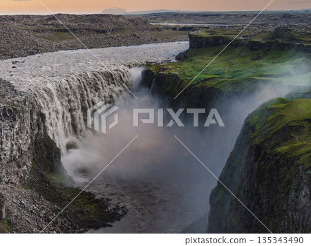Dettifoss waterfall cascades into a deep canyon with rugged cliffs and green moss. Mist rises from the falls under soft sunrise or sunset hues. 135343490