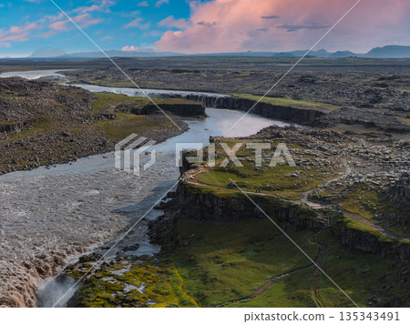 Dettifoss waterfall cascades over rugged cliffs in Iceland, surrounded by rocky terrain, green vegetation, and a winding path under a partly cloudy sky. Dettifoss waterfall cascades over rugged cliffs in Iceland, surrounded by rocky terrain, green vegetation, and a winding path under a partly cloudy sky. 135343491