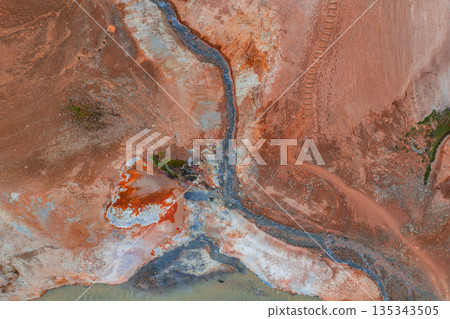 Aerial image of Iceland's geothermal terrain with red, orange, and white hues, a winding stream, visible mud pots, and tracks in the upper right. 135343505