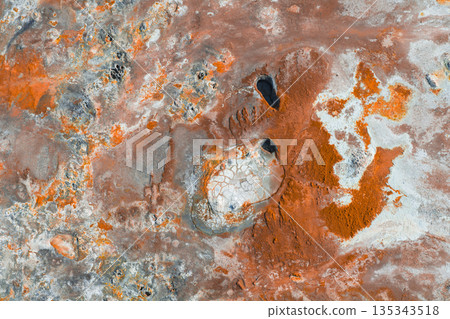 Geothermal terrain in Iceland with vibrant orange, red, and white hues, featuring two dark oval pools surrounded by cracked, pale surfaces. 135343518