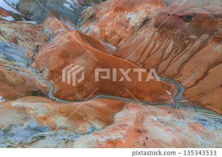 The image captures the vibrant orange and red rhyolite hills of Kerlingarfjoll, with steam vents, snow patches, and hikers on winding trails in Iceland. 135343533