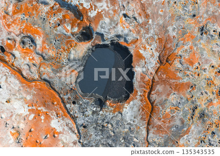 Aerial image of a geothermal area in Iceland featuring vibrant orange and gray hues, a dark oval pool, rugged textures, cracks, and small craters. 135343535