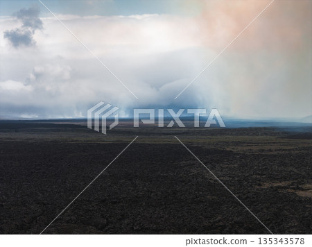 Dark volcanic terrain with cooled lava fields stretches into the distance under low clouds and mist, with hints of orange and blue in the sky. Dark volcanic terrain with cooled lava fields stretches into the distance under low clouds and mist, with hints of orange and blue in the sky. 135343578
