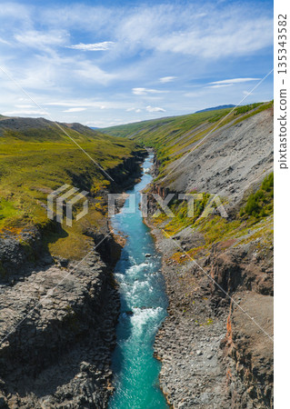 A vibrant turquoise river flows through Studlagil Canyon's basalt rock gorge, surrounded by green hills and rugged terrain under a clear blue sky. A vibrant turquoise river flows through Studlagil Canyon's basalt rock gorge, surrounded by green hills and rugged terrain under a clear blue sky. 135343582