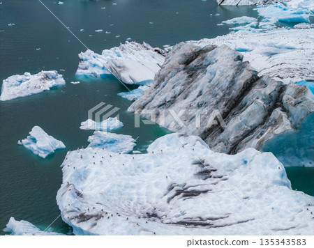 Aerial view of a glacial lagoon in Iceland with massive icebergs in turquoise water, volcanic ash streaks, and small birds on the ice. Aerial view of a glacial lagoon in Iceland with massive icebergs in turquoise water, volcanic ash streaks, and small birds on the ice. 135343583