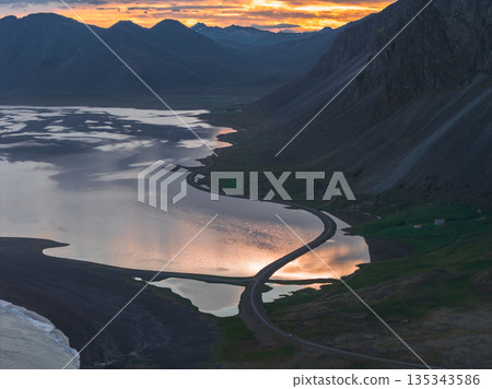 Aerial view of a coastal landscape in Iceland with a winding road, black sand beach, reflective water, rugged mountains, and a vibrant sunset. Aerial view of a coastal landscape in Iceland with a winding road, black sand beach, reflective water, rugged mountains, and a vibrant sunset. 135343586