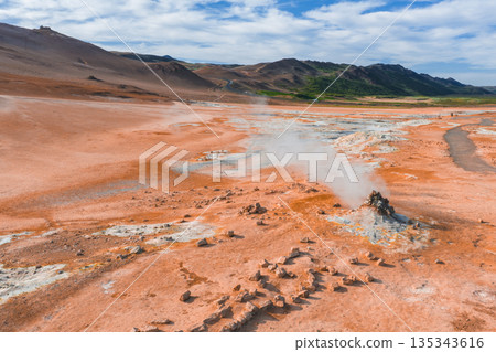 A geothermal landscape in Iceland featuring orange and red earth tones, steam from fumaroles, rocky terrain, and green hills under a blue sky. 135343616