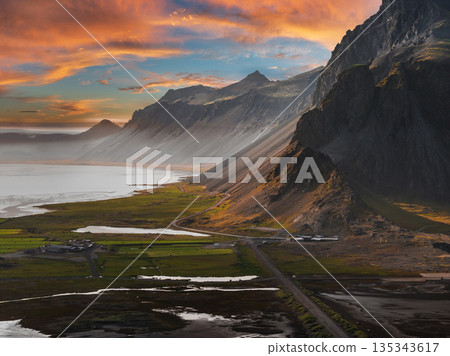 Aerial view of Stokknes Peninsula in Iceland featuring black sand beaches, rugged mountains, a winding road, and warm sunset hues over the landscape. Aerial view of Stokknes Peninsula in Iceland featuring black sand beaches, rugged mountains, a winding road, and warm sunset hues over the landscape. 135343617