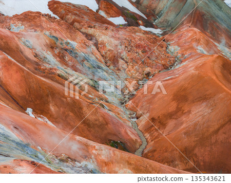 Vibrant orange, red, and brown rhyolite mountains with white snow patches and a stream cutting through rugged terrain in Landmannalaugar, Iceland. 135343621