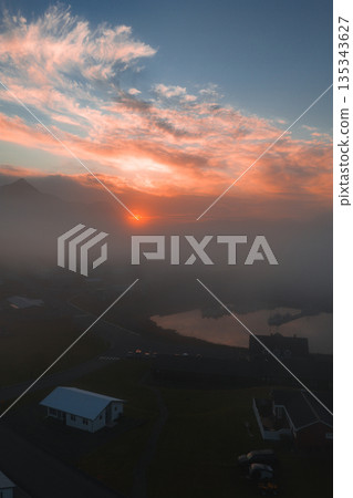 A serene sunset illuminates a misty Icelandic scene with a white roofed house, winding roads, grassy terrain, calm water, and fog covered mountains. 135343627