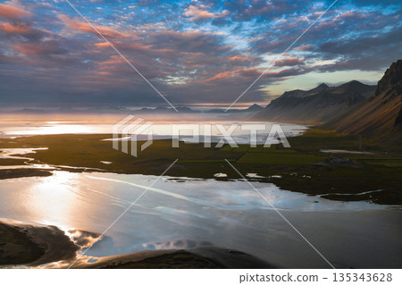 Aerial view of Iceland's Stokknes Peninsula at sunset, featuring reflective water channels, green fields, rugged mountain ridges, and a colorful sky. Aerial view of Iceland's Stokknes Peninsula at sunset, featuring reflective water channels, green fields, rugged mountain ridges, and a colorful sky. 135343628