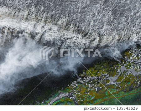 Dettifoss waterfall cascades with immense force, surrounded by mist, rocky terrain, green moss, and mineral rich water in Iceland's rugged landscape. 135343629