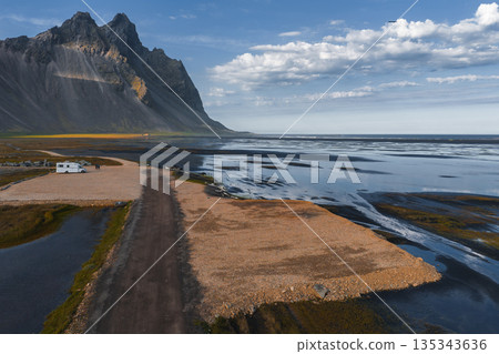 The Stokknes Peninsula features the Vestrahorn mountains, black sand beaches, shallow tidal waters, a gravel road, and a camper van under a blue sky. The Stokknes Peninsula features the Vestrahorn mountains, black sand beaches, shallow tidal waters, a gravel road, and a camper van under a blue sky. 135343636