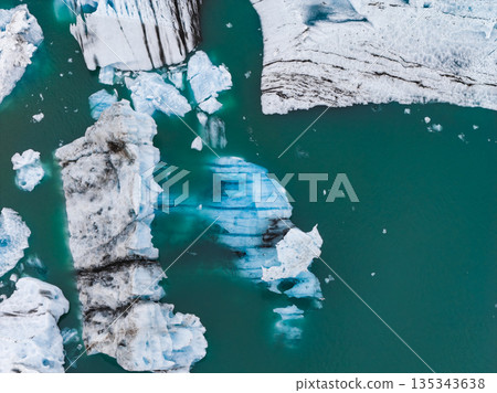 Icebergs with black volcanic ash and blue ice float in a turquoise glacial lagoon. The fractured shapes and vibrant water highlight Iceland's geology. 135343638