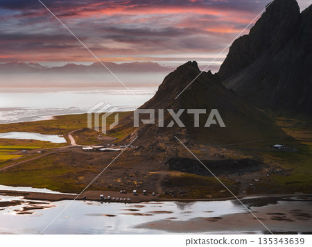 The Stokksnes Peninsula in Iceland features a triangular mountain, reflective water pools, and a vibrant sunset sky transitioning from orange to purple. 135343639