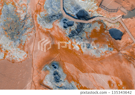 Geothermal area in Iceland with vibrant orange and blue hues, steaming vents, bubbling mud pots, and a wooden walkway for visitors. 135343642