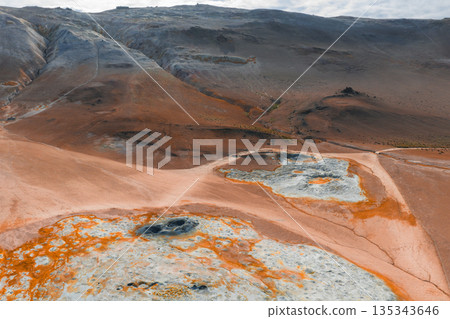 Vibrant orange, red, and white mineral deposits surround steaming mud pots and fumaroles, set against rugged hills in a barren Icelandic landscape. 135343646