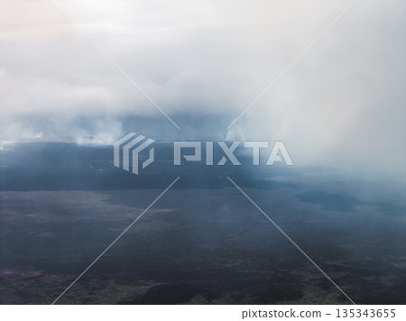 Aerial view of a rugged volcanic terrain in Iceland with steam rising from the ground. Dark lava fields and haze create a dramatic atmosphere. 135343655