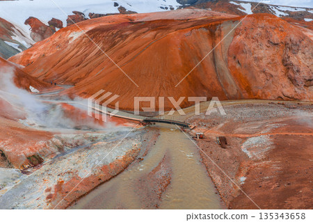 Kerlingarfjoll's vibrant geothermal area features orange rhyolite mountains, a steaming river with a wooden bridge, and snow capped peaks in the distance. Kerlingarfjoll's vibrant geothermal area features orange rhyolite mountains, a steaming river with a wooden bridge, and snow capped peaks in the distance. 135343658