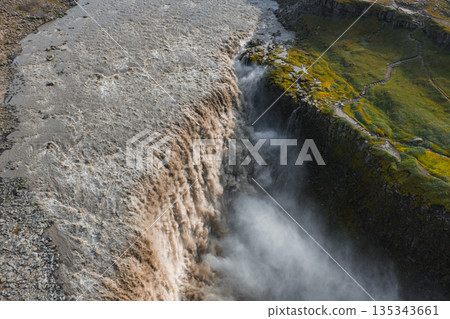 Dettifoss waterfall cascades powerfully into a misty canyon, with muddy waters and lush green terrain contrasting rugged rocky surroundings. 135343661