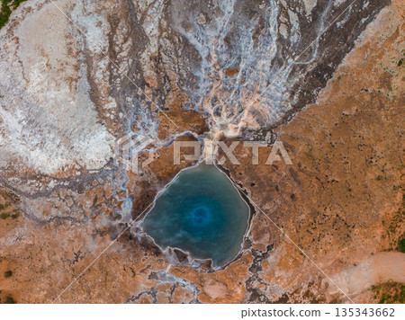 Geothermal hot spring in Iceland with a vivid blue pool, surrounded by multicolored mineral deposits, steaming fissures, and intricate geological patterns. 135343662