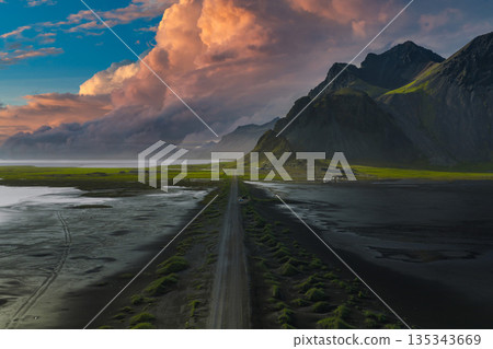 A long road stretches through black sand terrain toward rugged mountains on Iceland's Stokknes Peninsula, illuminated by a vibrant sunset. A long road stretches through black sand terrain toward rugged mountains on Iceland's Stokknes Peninsula, illuminated by a vibrant sunset. 135343669