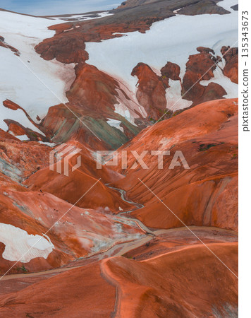 Vibrant rhyolite mountains in Landmannalaugar, Iceland, display red, orange, and brown hues with snow patches and a winding trail through rugged terrain. 135343673