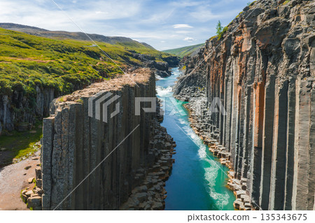 Studlagil Canyon in Iceland features dramatic basalt columns, a vibrant turquoise river, lush green landscapes, and rolling hills under a blue sky. Studlagil Canyon in Iceland features dramatic basalt columns, a vibrant turquoise river, lush green landscapes, and rolling hills under a blue sky. 135343675