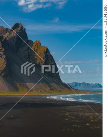 The Stokksnes Peninsula features the jagged Vestrahorn mountain, a black sand beach, gentle waves, sparse green vegetation, and distant mountain ranges. The Stokksnes Peninsula features the jagged Vestrahorn mountain, a black sand beach, gentle waves, sparse green vegetation, and distant mountain ranges. 135343680